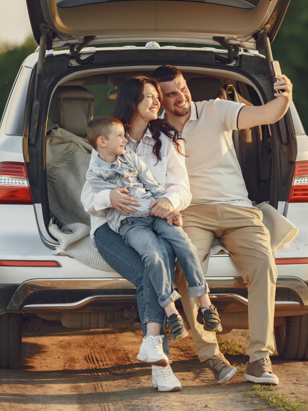 Um casal, homem, mulher e uma criança, sentados no portal malas do carro, do lado de fora do veículo, tirando uma foto juntos.